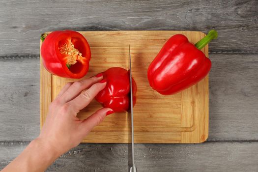 Tabletop view, woman hand holding red bell pepper, cutting it with chef knife on chopping board.