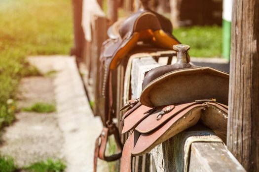 Old horse saddle placed on wooden fence next to house, lit by sun.