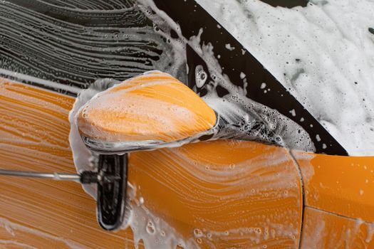 Yellow car washed in self serve carwash, brush leaving strokes on side, mirror covered with shampoo and foam.