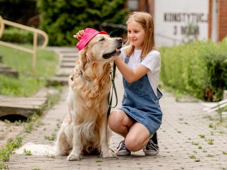Preteen girl wearing hat hugging golden retriever dog sitting outdoors in summertime. Pretty kid petting fluffy doggy pet in city