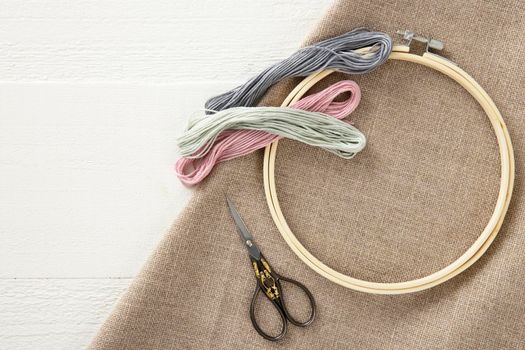 Embroidery floss, vintage style scissors, wooden hoop and cotton cloth on painted wooden surface with copy space, flat lay