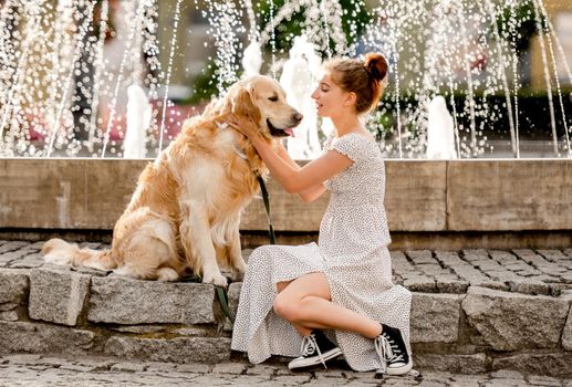 Girl in beautiful dress petting golden retriever dog at street in summer day close to fountain. Pretty teenager with purebred doggy pet in city with ancient architecture