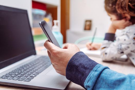 detail of the hand of a man working at home with his laptop holding a mobile phone while his daughter is busy drawing, work at home concept due to the coronavirus pandemic