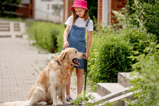 Preteen girl wearing hat with golden retriever dog outdoors in summertime. Pretty kid with fluffy doggy pet walking in city