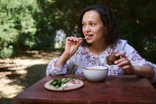 Charming woman sits at rustic dining table, tastes Ukrainian national dish- traditional Borscht with green onion, sliced bacon, parsley and whole grain rye bread, enjoying lunch in the countryside
