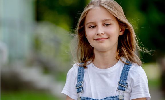 Beautiful preteen girl in city in summertime closeup portrait. Pretty female kid looking at camera in sunny day with blurred background