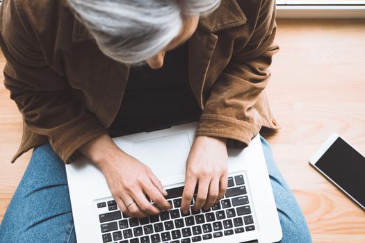 Grey haired woman typing laptop keyboard sitting on wooden windowsill with mobile phone on it. Top view. Selective focus on female hands. 