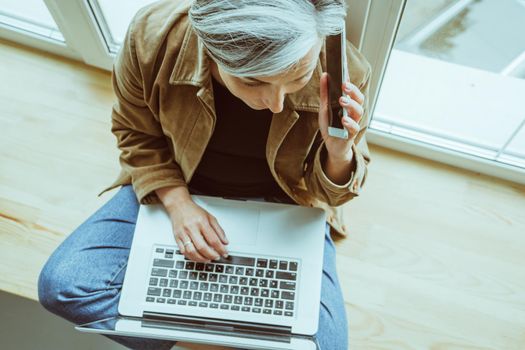 Mature woman talks mobile phone using laptop computer. Gray haired Caucasian woman in casual works sitting crossed legs on window sill. High angle view.