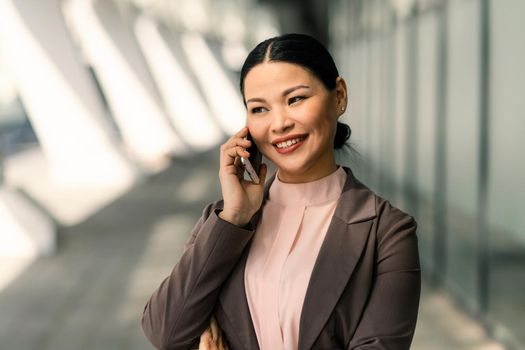 Smiling Asian businesswoman in suit talking on mobile phone against backdrop of business center. High quality photo.