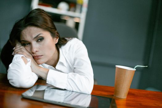 Tired or bored business woman relax at workplace. Young Caucasian woman rest at office table with coffee paper cup and digital tablet on it. Crisis concept. Tinted image.