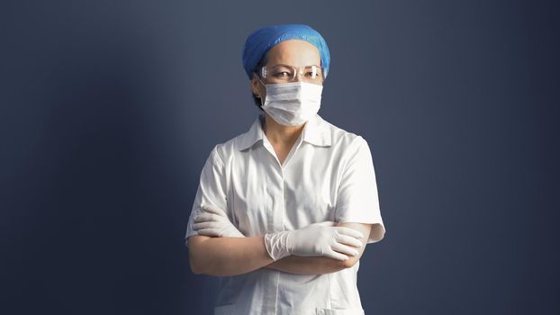 Female doctor looks at camera crossed arms. Female medic in mask and white uniform, portrait on gray background.