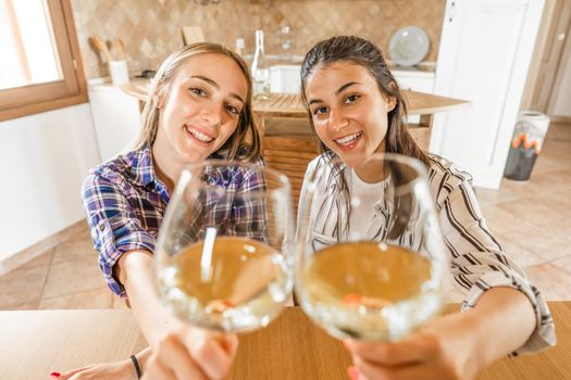 POV portrait of two girls looking at camera holding champagne glasses in selective focus. Students celebrating online with alcohol using video conferencing technology. New normal social activity