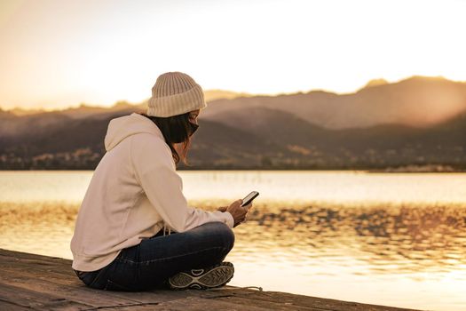 Unrecognizable young woman in white wool hat sitting on a pier alone wearing protective face mask using smartphone to keep in touch with friends and family due to Coronavirus social distancing