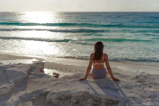 A woman in a striped swimsuit sits on a rock and looks out at the sea. Mexico.