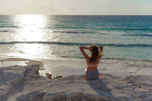 A woman in a striped swimsuit sits on a rock and looks out at the sea. Mexico.