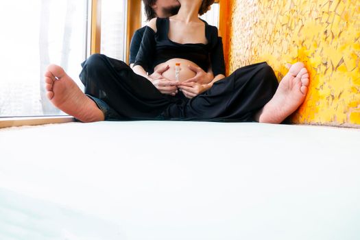 pregnancy, love, family and people concept - pregnant woman and her husband making hand heart gesture on her belly sitting in the lotus position. High quality photo