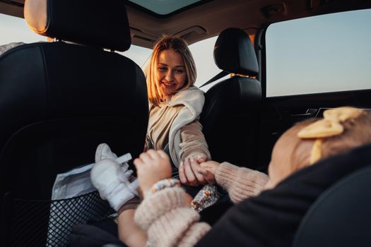 Young Mother Holding Her Baby Daughter by Hand While She Sitting in Child Car Seat in Car, Young Family on Road Trip