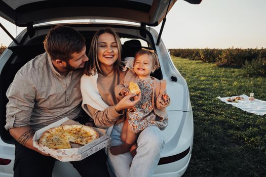 Happy Young Family Sitting in Trunk of SUV Car and Eating Pizza, Mom Dad and Baby Daughter Having Fun Time on Weekend Outdoors