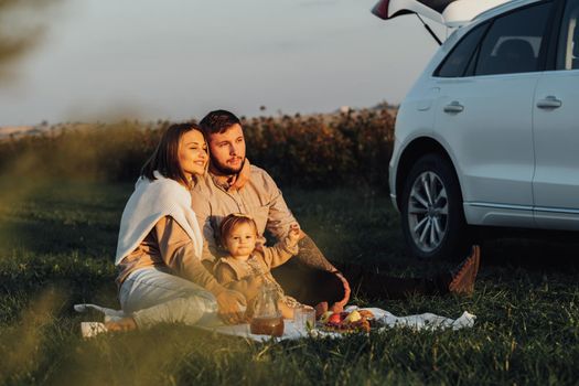 Happy Young Family Enjoying Road Trip on SUV Car, Mom Dad and Baby Daughter Having Picnic on Weekend Outdoors at Sunset