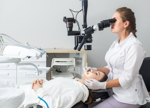 Female dentist using dental microscope treating patient teeth at dental clinic office. Medicine, dentistry and health care concept