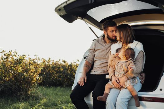 Young Caucasian Family Sitting in Trunk of SUV Car, Mom and Dad with Baby Daughter Having Fun Time on Weekend Outdoors