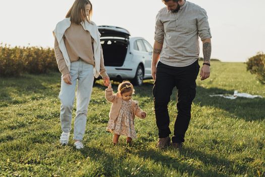 Young Family Enjoying Road Trip on SUV Car, Mother and Tattooed Father with Baby Daughter Walking Outdoors in Field at Sunset