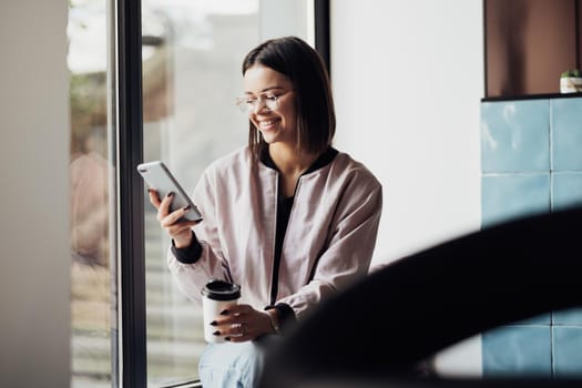 Young Woman in Glasses Drinking Coffee and Using Smartphone While Sitting on Windowsill