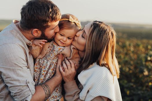 Close Up Portrait of Happy Young Caucasian Family Outdoors, Mother and Father Kissing Their Little Baby Daughter at Sunset