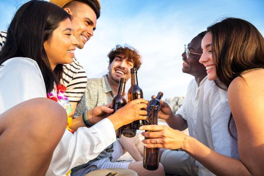 Close up of happy, diverse group of friends toasting with beer together at the beach. Multiracial young people having fun outdoor. Vacation and friendship concept.