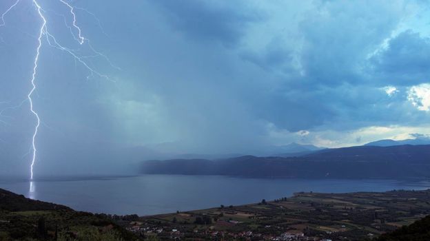 A single bright bolt of lightning captured and reflected in Lake Trichonida, the largest natural lake in Greece. Dark blue storm clouds roll in over mountains in the distance accompanied by rain.