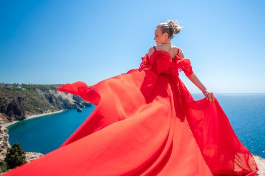 Blonde with long hair on a sunny seashore in a red flowing dress, back view, silk fabric waving in the wind. Against the backdrop of the blue sky and mountains on the seashore