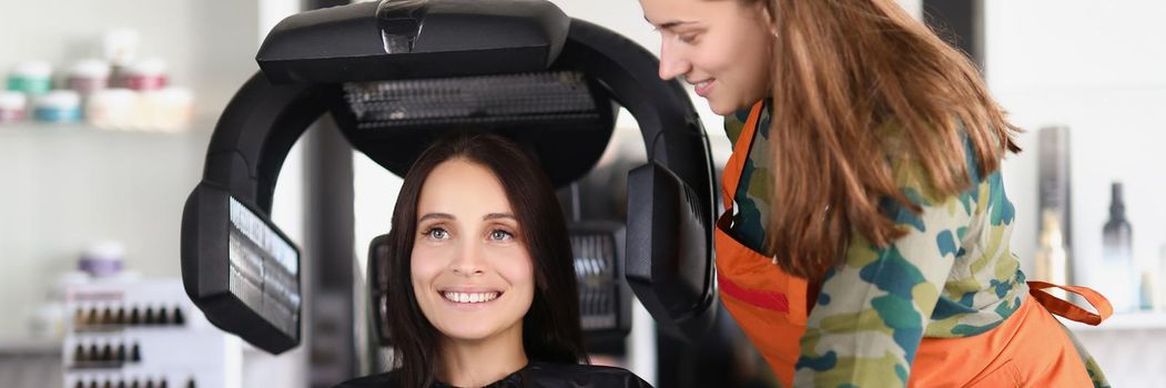 Portrait of young lady under hairdressing machine while looking in mirror. Visit master for beautiful look and good hair quality. Self care, beauty concept