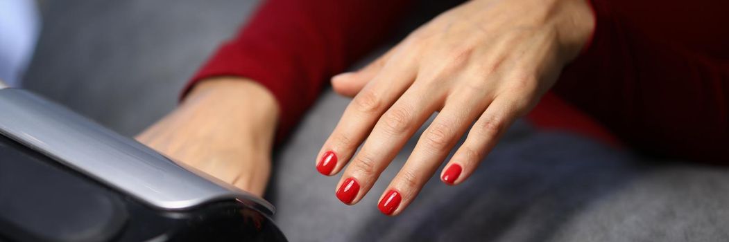 Close-up of woman client putting hand with fresh red nails under lamp to dry it. Spend time on beauty procedure in luxury studio. Beauty, wellness concept