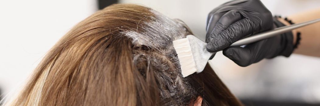 Close-up of hair master apply dye colour on hair with grey brush tool. Female client enjoying beauty procedure. Wellness, beauty salon, self care concept