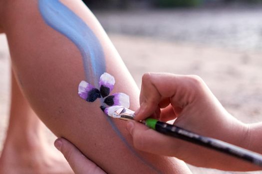 Close up view of the hand of an artist painting a flower in the leg of a woman in the beach