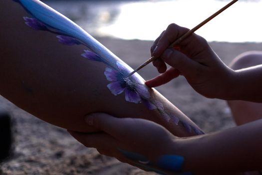 Close up view of the hand of an artist painting the body of a woman during sunset on a beach