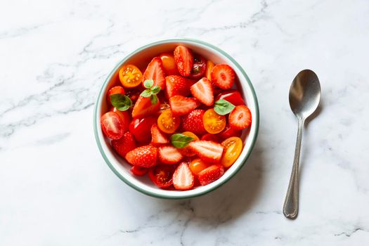 portion of strawberry and tomato cherry salad decorated with basil leaves, marble background, top view