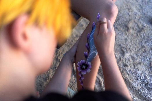 Top view and close up photo of an artist painting a flower in the leg of a woman on the beach