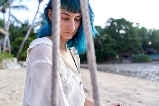 Alternative woman with blue hair sitting on a swing on a tropical beach