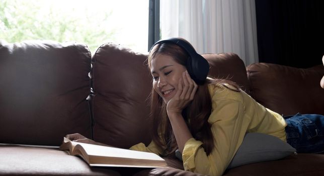Beautiful young asian woman using laptop and wearing headphones listening to music and lying down on sofa in living room.