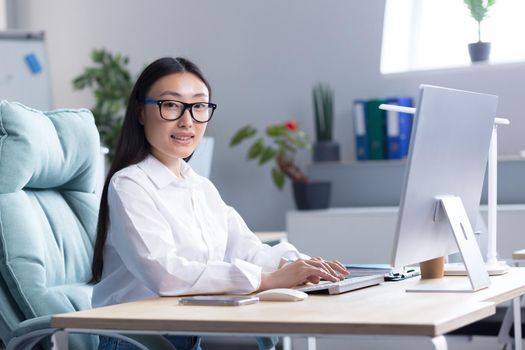 Portrait of a young Asian woman in the office. Businesswoman, director, freelancer, manager, sitting in a white shirt and glasses at the desk, working at the computer. He looks at the camera, smiles.