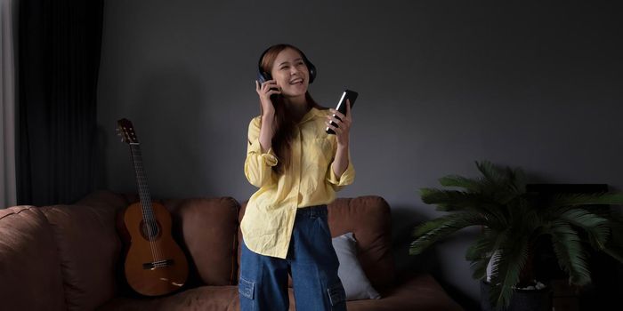 Young asian woman listening to music on couch in living room at home. Happy asia female using mobile smartphone, wearing headset and sitting on sofa.
