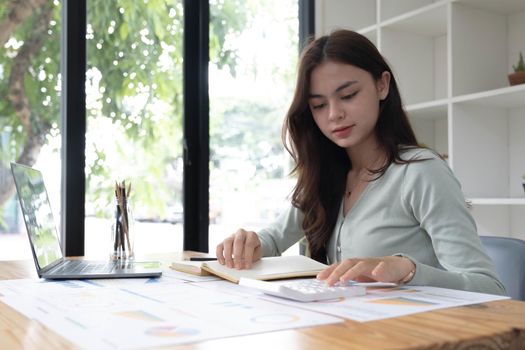 Portrait of asian business woman paying bills online with laptop in office. Beautiful girl with computer and chequebook, happy paying bills. Startup business financial calculate account concept.