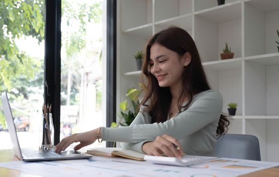 Portrait of asian business woman paying bills online with laptop in office. Beautiful girl with computer and chequebook, happy paying bills. Startup business financial calculate account concept.