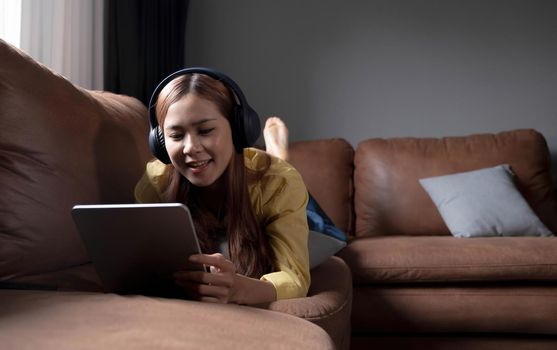 Beautiful young asian woman using laptop and wearing headphones listening to music and lying down on sofa in living room.