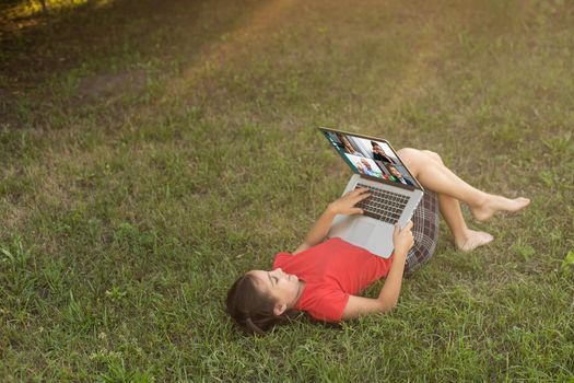 Beautiful young lady is chatting with friends outdoors as it is very useful to be in nature. Laptop is the best friend for a child nowadays. Green and vast meadow.