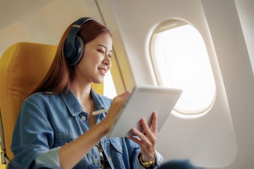 Attractive portrait of an Asian woman sitting in a window seat in economy class using tablet and listening to instrumental music during an airplane flight, travel concept, vacation, relaxation.