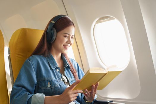 Attractive portrait of an Asian woman sitting in a window seat in economy class reading a book and listening to instrumental music during an airplane flight, travel concept, vacation, relaxation.