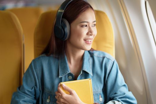 Attractive portrait of an Asian woman sitting in a window seat in economy class reading a book and listening to instrumental music during an airplane flight, travel concept, vacation, relaxation.