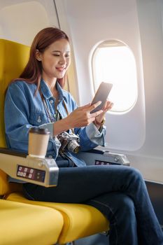 Attractive portrait of Asian woman sitting at window seat in economy class using mobile phone during inflight, travel concept, vacation, relax.
